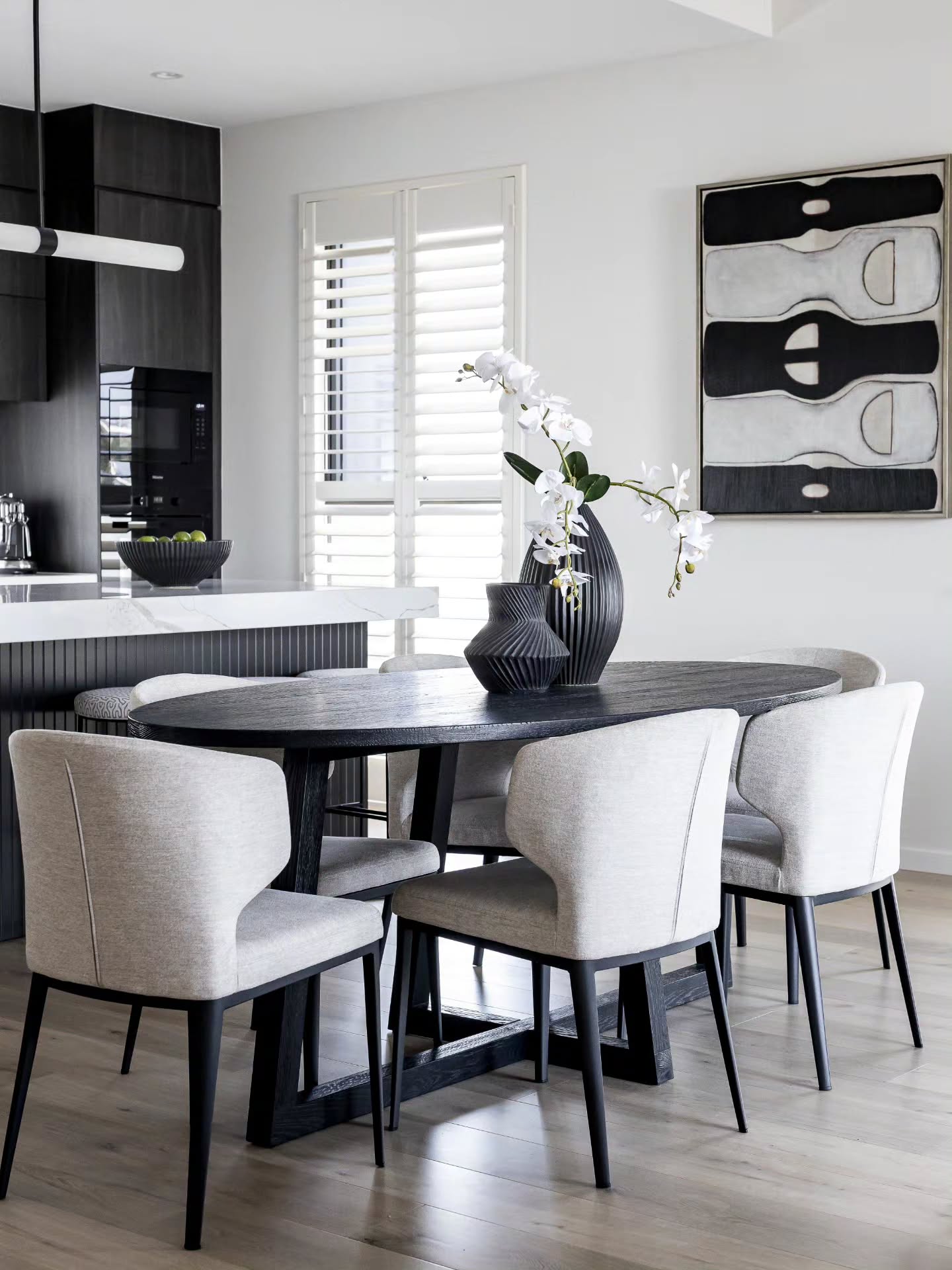 Oval dining table with pale upholstered chairs and orchids below abstract black-and-white artwork — Monochrome Residence