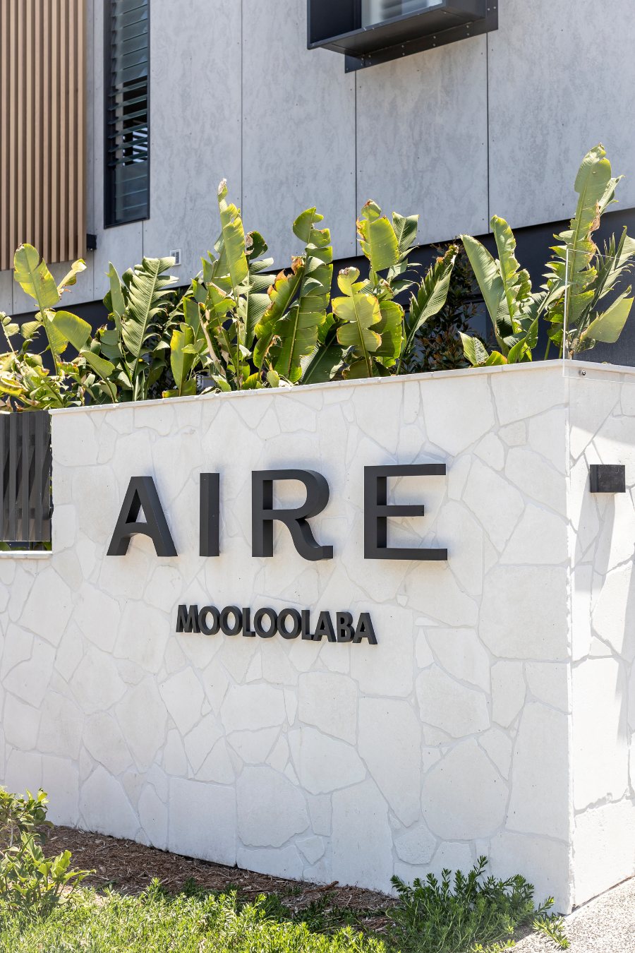Black "AIRE Mooloolaba" building signage on a stone-clad wall with banana palms — Monochrome Residence