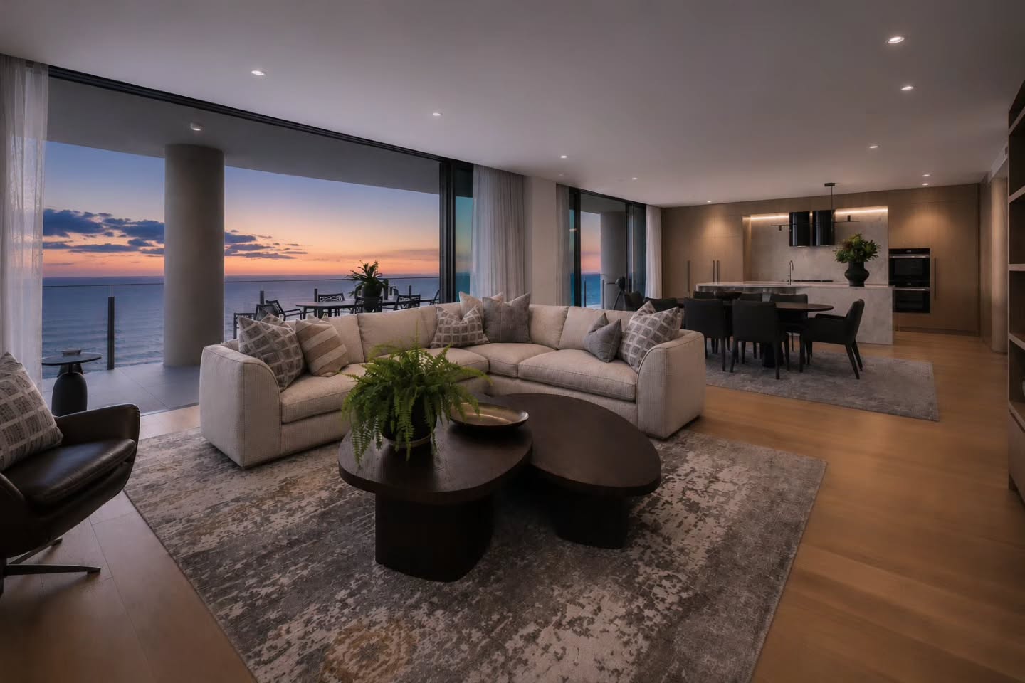 Open-plan living opening to an ocean-view terrace at sunset, cream sectional and dining beyond — Oceanfront Apartment